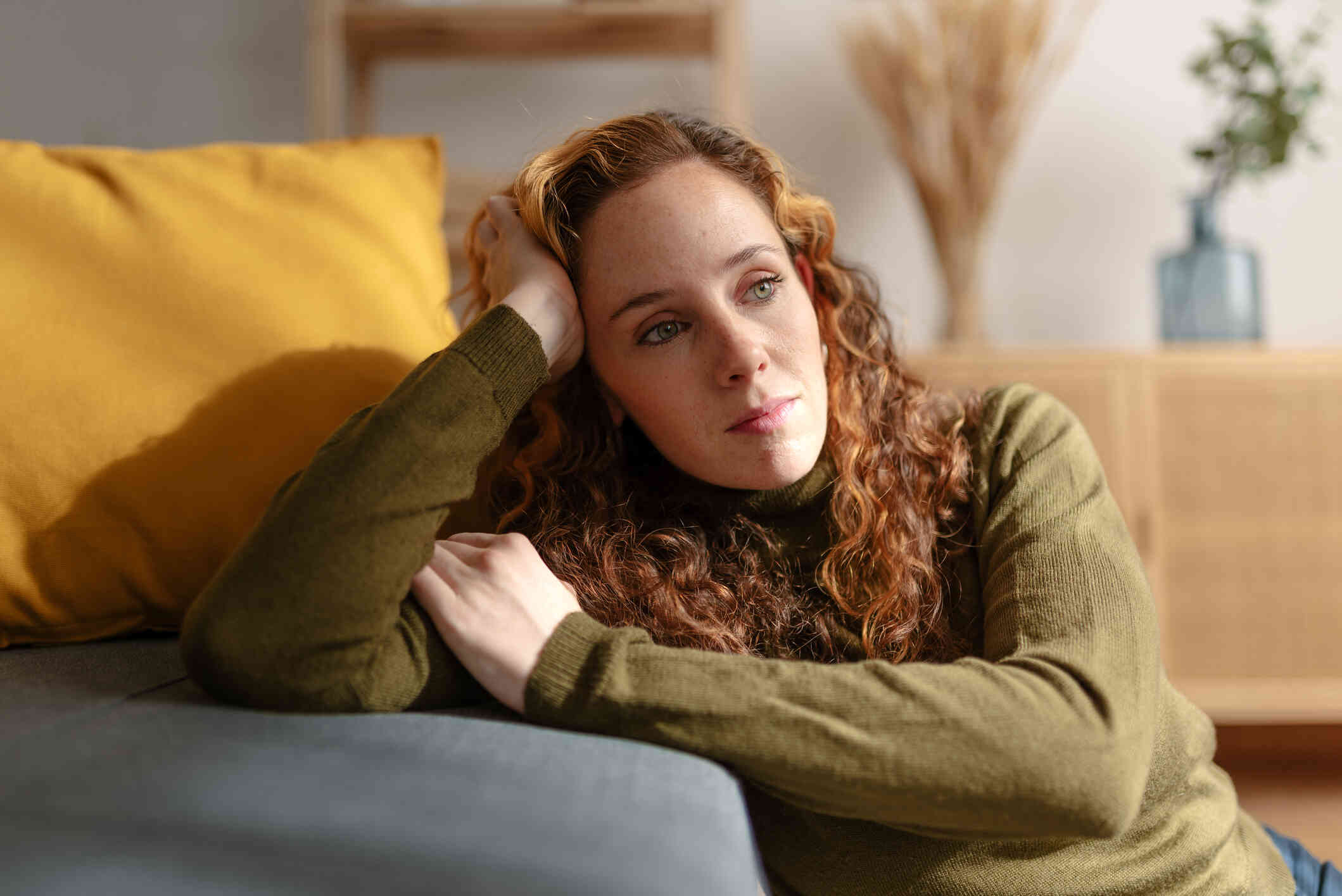 A woman in a green turtleneck sits on the living room floor and leans against the couch with her head in her hand as she gazes off sadly.